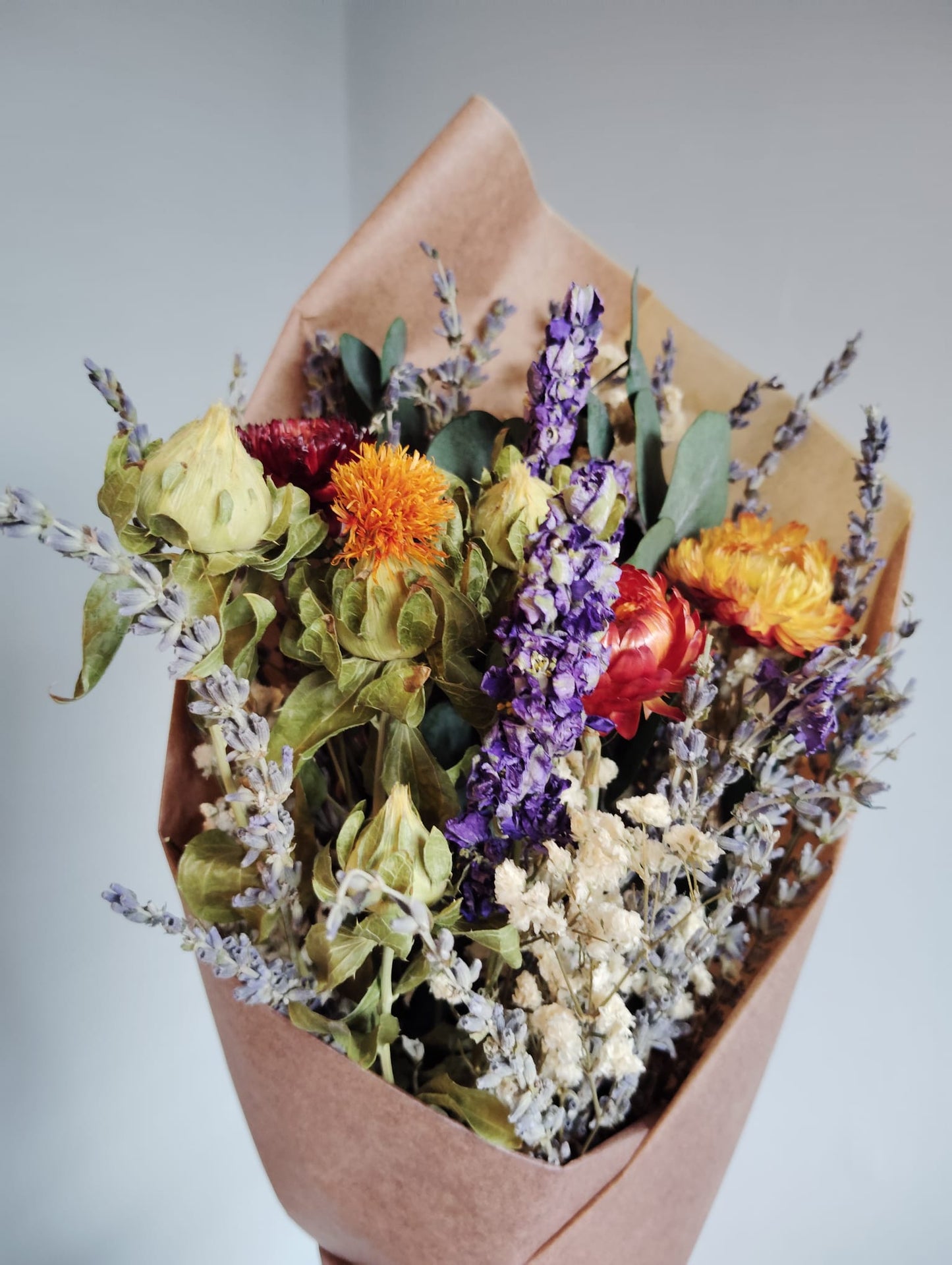 A rustic dried flower bouquet featuring fragrant purple lavender stems, bright orange safflowers, and preserved eucalyptus wrapped in brown kraft paper.